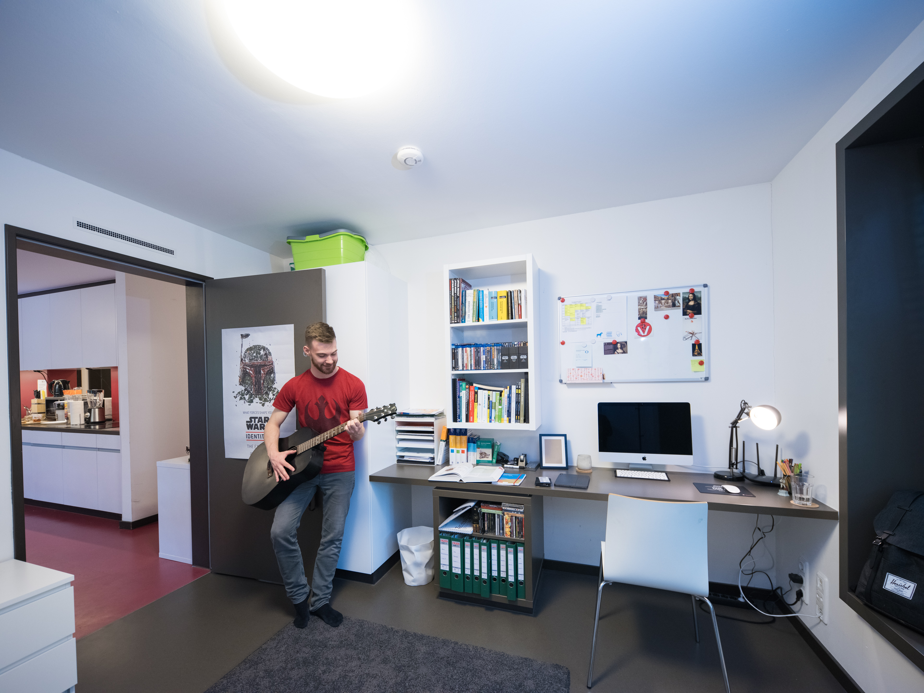 Junger Mann in rotem T-Shirt spielt auf seiner Gitarre, gelehnt an eine offene Tür, man sieht einen aufgeräumten Schreibtisch und im Hintergrund eine Küche.  